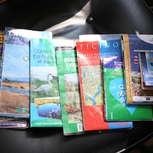 Peut inclure: Une collection de brochures de voyage et de cartes touristiques pliées présentant divers paysages, un cygne, un papillon et du texte incluant Courmayeur, Ticino et Mikroregion Nove Hamry. Les guides en papier colorés sont alignés.