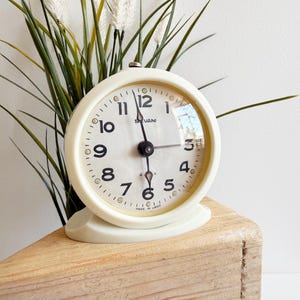 Può includere: Una sveglia vintage color crema con quadrante rotondo e numeri neri. Il quadrante dell'orologio indica "SVANI" e "MADE IN USSR". L'orologio è posto su un blocco di legno, con piante verdi e bianche sullo sfondo.