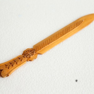 May include: A decorative wooden letter opener with a detailed floral and leaf design on the handle. The blade has a textured pattern. The opener is a light brown colour.