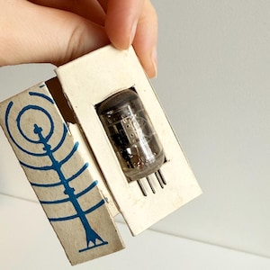 May include: A vintage vacuum tube in its original cardboard packaging. The tube is clear with internal components visible. The box is off-white with a blue graphic design on the side.