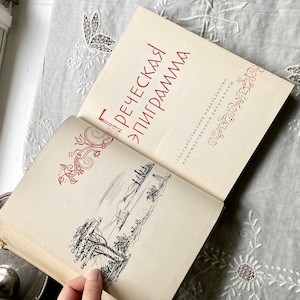 Può includere: Un libro vintage aperto con copertina e pagine color crema. La copertina presenta testo rosso ed elementi decorativi. Le pagine aperte rivelano un'illustrazione di paesaggio in bianco e nero. Il libro è su una superficie ricamata bianca.