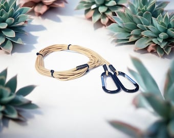 May include: A beige rope leash with black carabiner clips. The leash is coiled and secured with black ties. The background features green and pink succulents on a white surface.