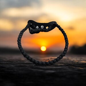 May include: A black slingshot with a braided cord forms a circle against a sunset backdrop. The slingshot's pouch is made of black material with silver rivets. The background features a vibrant orange and yellow sky.