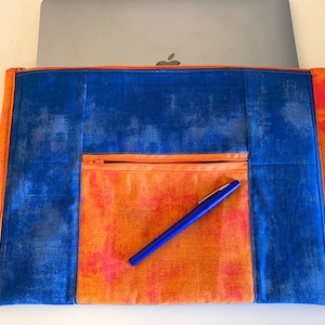 May include: A blue and orange laptop sleeve with a zippered pocket and a blue pen inside. The sleeve is made of a soft, textured fabric.