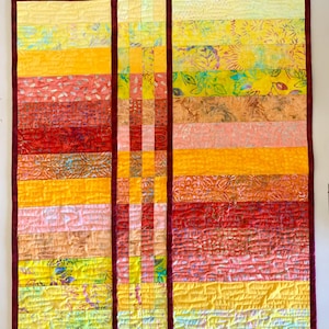 May include: A rectangular quilt featuring a gradient of warm colors including yellow, orange, and red. The design consists of horizontal fabric strips separated by a vertical column of small squares, all bordered by a dark maroon fabric binding.