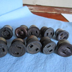 May include: A collection of vintage electrical sockets with a dark brown and black color scheme. The sockets have various designs and markings, including the brand name "ARROW". They are arranged on a light blue surface.