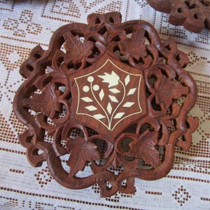 Puede incluir: Un posavasos redondo de madera tallada intrincadamente con un diseño floral. El posavasos es de color marrón intenso, con un diseño central incrustado en blanco. El diseño presenta un patrón estilizado de flores y hojas, rodeado de hojas y vides talladas.