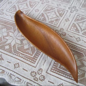 May include: A long, leaf-shaped wooden bowl with a smooth, polished surface. The bowl is a warm brown color and appears to be hand-carved. It rests on a white, patterned tablecloth.