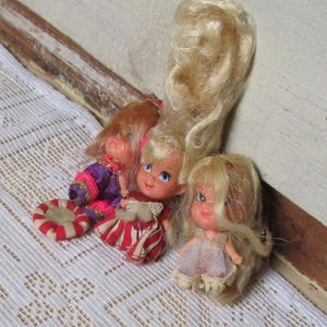 Könnte beinhalten: Drei Vintage-Puppen mit langen blonden Haaren. Die Puppen tragen bunte Kleider und haben bemalte Gesichter. Die Puppen sitzen auf einer weißen Spitzentischdecke.
