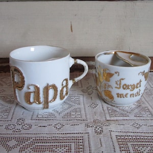 May include: Two white ceramic mugs with gold lettering. One mug says "Papa" and the other says "Forget me not".