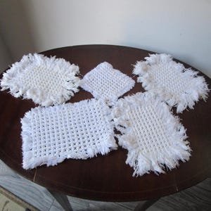 Puede incluir: Cinco posavasos blancos de ganchillo con bordes con flecos están colocados sobre una mesa de madera marrón oscuro. Los posavasos son de varias formas y tamaños, incluyendo diseños cuadrados y rectangulares.