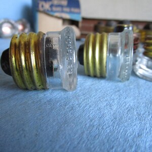 Può includere: Primo piano di diversi portalampada vintage in vetro e ottone. Le basi hanno un corpo in vetro trasparente con una base a vite in metallo color ottone. Le basi sono disposte su una superficie azzurra.