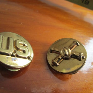 May include: Two gold-colored metal buttons. One button features the letters "US" in raised lettering. The other button features a crossed cannon design.