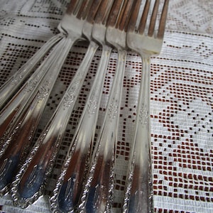 May include: A set of silver forks with ornate designs on the handles. The forks are arranged in a fan shape on a white lace tablecloth.