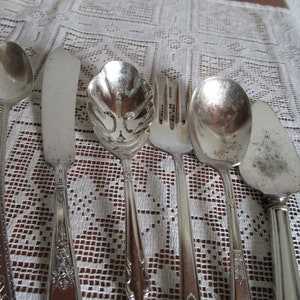 Puede incluir: Un juego de seis cubiertos plateados, que incluye un cuchillo para mantequilla, una cuchara para servir, una cuchara para sopa, un tenedor, un cuchillo para pastel y una cuchara ranurada. Los cubiertos están dispuestos sobre un mantel de encaje blanco.