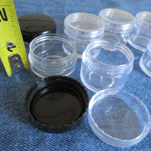 Op de afbeelding: Een set van acht doorzichtige plastic containers met zwarte deksels. De containers zijn ongeveer 2,5 cm in diameter en 2,5 cm hoog. Een gele meetlint is op de achtergrond.