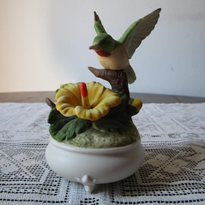 May include: A ceramic figurine of a hummingbird perched on a branch above a yellow hibiscus flower. The hummingbird has green and white wings, a red throat, and a long beak. The flower sits atop a white, rounded base.