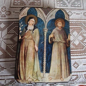 May include: Rectangular religious art print featuring two figures with halos, set against a blue backdrop with architectural details. The figures are dressed in robes, one holding a flower. The artwork has a vintage, aged appearance.