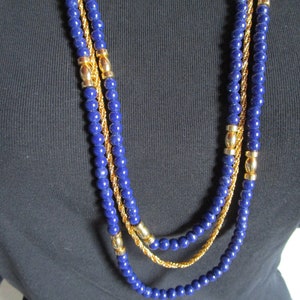Puede incluir: Un collar de tres capas con tres hileras de cuentas de color azul oscuro y detalles de cadena dorada.
