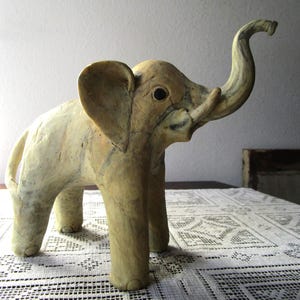 Può includere: Una statuetta di elefante artigianale di colore chiaro con una proboscide sollevata e grandi orecchie. La scultura ha una superficie strutturata ed è esposta su una tovaglia di pizzo bianco. Le zanne dell'elefante sono visibili e gli occhi sono scuri.