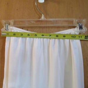 May include: White slip with a gathered elastic waistband. The slip is measured with a tape measure, showing a length of 11 inches.