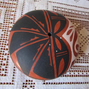 Puede incluir: Objeto cerámico redondo pintado a mano con un agujero central. El diseño presenta un fondo negro con líneas rojo-naranja radiantes y formas de hojas. Una sección del objeto es de color beige claro con patrones decorativos rojo-naranja y blancos.