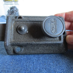 Puede incluir: Un pestillo de puerta Yale vintage de color gris oscuro. El pestillo tiene una perilla redonda con el logotipo de Yale y un botón pequeño y redondo. El metal tiene un aspecto texturizado. El pestillo está diseñado para la seguridad y probablemente para una puerta.