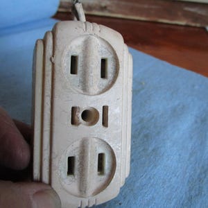 Puede incluir: Un adaptador de toma de corriente vintage blanco con dos tomas estándar y una toma redonda en el centro.