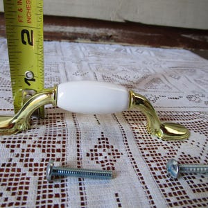 May include: A cabinet pull with a white ceramic centre and gold-toned metal accents. The pull is approximately 10 cm long, with mounting screws included. The hardware is shown on a white lace tablecloth.