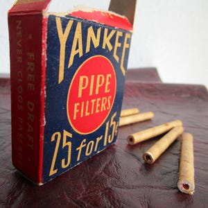 May include: A vintage box of Yankee Pipe Filters, blue with red and gold lettering. The box reads "Yankee Pipe Filters 25 for 15c" and "Free Draft, Never Clogs Passage".  The box is next to a pile of five brown pipe filters.