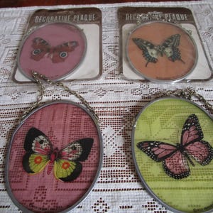 May include: Four decorative plaques with butterfly designs. Two plaques are pink with black butterflies, one is green with a pink butterfly, and one is orange with a black and white butterfly. Each plaque has a silver chain for hanging.