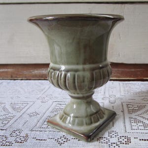 Pode incluir: Uma urna decorativa de cerâmica vidrada em tons de verde e castanho. A urna tem um topo alargado, uma secção média canelada e uma base quadrada. Mede aproximadamente 20 cm de altura.