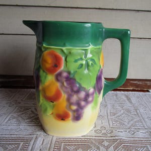 May include: A ceramic pitcher with a green handle and rim, featuring a relief design of peaches, grapes, and leaves. The pitcher has a cream-colored base and is likely used for serving beverages. The pitcher is a decorative item.