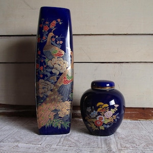 Puede incluir: Dos jarrones de cerámica. Un jarrón alto y rectangular de color azul cobalto con un diseño de pavo real y flores. Un tarro más pequeño y redondo de color azul cobalto con tapa y diseño floral. Ambos tienen detalles dorados.