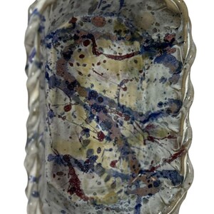 May include: An oval ceramic bowl with a wavy, scalloped rim. The interior features a cream-colored base glaze decorated with an abstract splatter pattern in shades of deep blue, dark red, and muted brown, creating a textured, multi-colored effect.