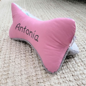 Könnte beinhalten: Ein rosa-graues knochenförmiges Kissen mit dem Namen "Antonia" bestickt.
