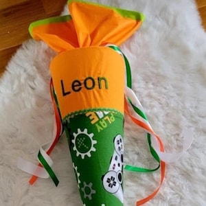 Könnte beinhalten: Ein grüner und orangefarbener, kegelförmiger Schultüte mit dem Namen "Leon" auf der orangenen Seite bestickt. Die grüne Seite hat ein Videospiel-Controller-Design und die Worte "Play Game" darauf geschrieben. Die Tüte ist mit weißen, grünen und orangenen Bändern verziert.