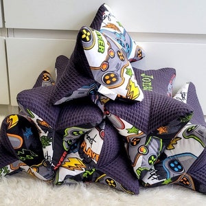 May include: A stack of six black and white gaming themed pillows with a purple back. The top pillow has the word "Gamer" embroidered on it. The pillow below has the name "John" embroidered on it.