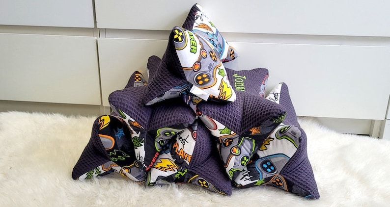 May include: A stack of six black and white gaming themed pillows with a purple back. The top pillow has the word "Gamer" embroidered on it. The pillow below has the name "John" embroidered on it.