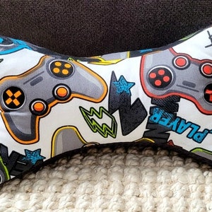 May include: A white and gray bone-shaped pillow with a gamer theme. The pillow is covered in a fabric with a pattern of video game controllers, lightning bolts, and the word "Player".