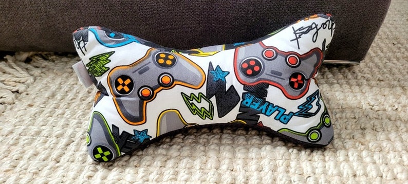 May include: A white and gray bone-shaped pillow with a gamer theme. The pillow is covered in a fabric with a pattern of video game controllers, lightning bolts, and the word "Player".