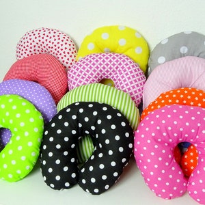 May include: A collection of colorful travel neck pillows in various patterns. Pillows include polka dots, hearts, stripes, and geometric designs in shades of pink, green, orange, yellow, and black. The pillows are U-shaped for neck support.