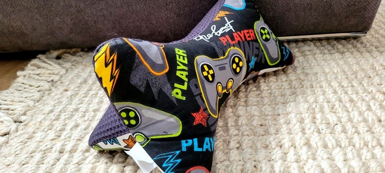May include: A black and gray neck pillow with a gamer theme. The pillow has a repeating pattern of video game controllers, lightning bolts, and the word "Player".