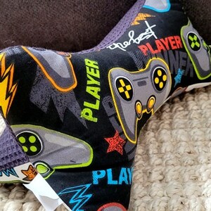 May include: A black and gray neck pillow with a gamer theme. The pillow is covered in a fabric with a repeating pattern of video game controllers, lightning bolts, and the words "Player" and "The Best".