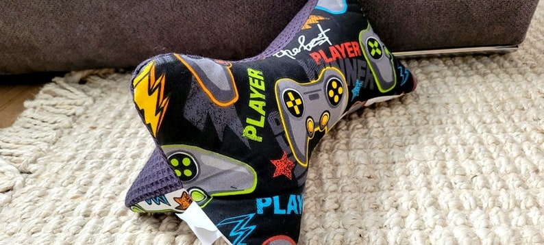 May include: A black and gray neck pillow with a gamer theme. The pillow is covered in a fabric with a repeating pattern of video game controllers, lightning bolts, and the words "Player" and "The Best".