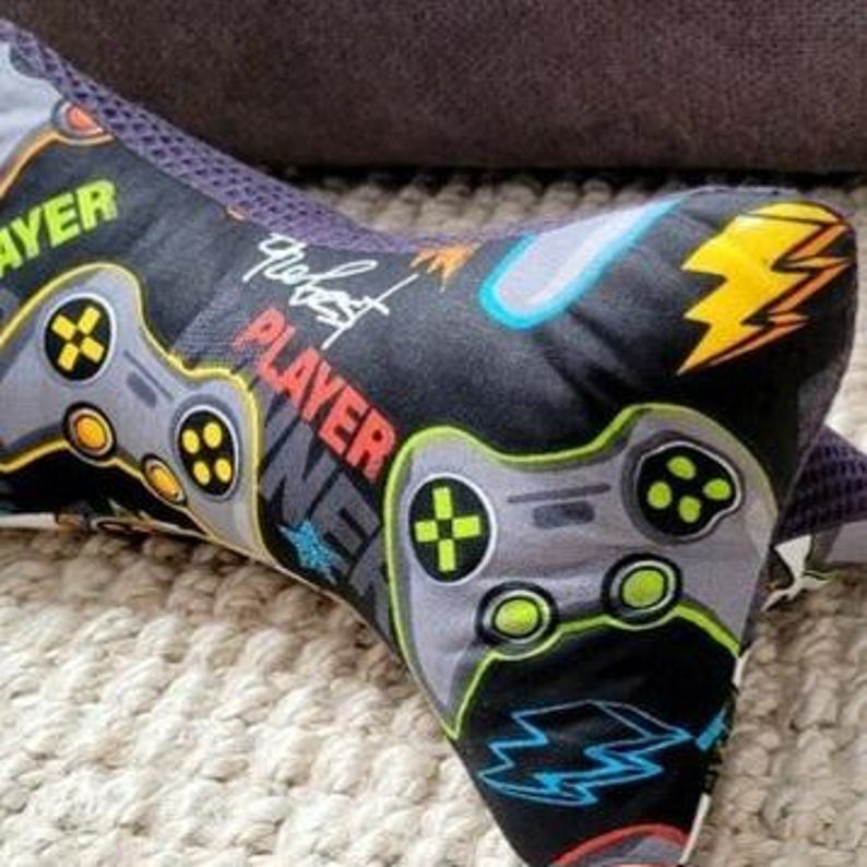 May include: A black and gray gaming themed neck pillow with the text "the best player" printed on it. The pillow has a mesh fabric on the back and a soft, plush fabric on the front. The pillow is decorated with images of game controllers, lightning bolts, and other gaming-related graphics.