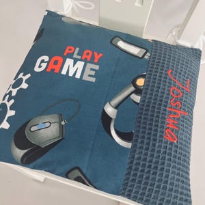 Könnte beinhalten: Ein quadratisches blaues Kissen mit einem Gaming-Motiv aus Computermäusen, Headsets und Zahnrad-Symbolen. Der Text "PLAY GAME" ist in Orange und Weiß aufgedruckt. Ein dunkelblauer Waffelstoff-Einsatz auf der rechten Seite zeigt den in Rot gestickten Namen "Joshua".