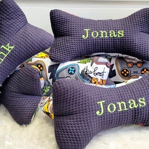 May include: Two dark purple bone-shaped pillows with the name "Jonas" embroidered on them. One pillow is on top of the other. A third pillow with the name "Dominik" embroidered on it is in the background.