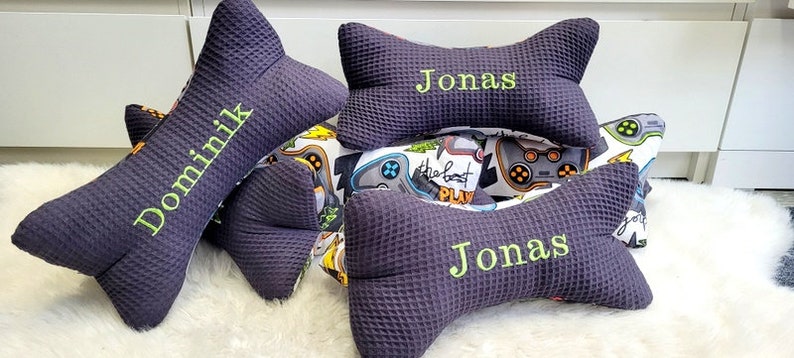 May include: Two dark purple bone-shaped pillows with the name "Jonas" embroidered on them. One pillow is on top of the other. A third pillow with the name "Dominik" embroidered on it is in the background.
