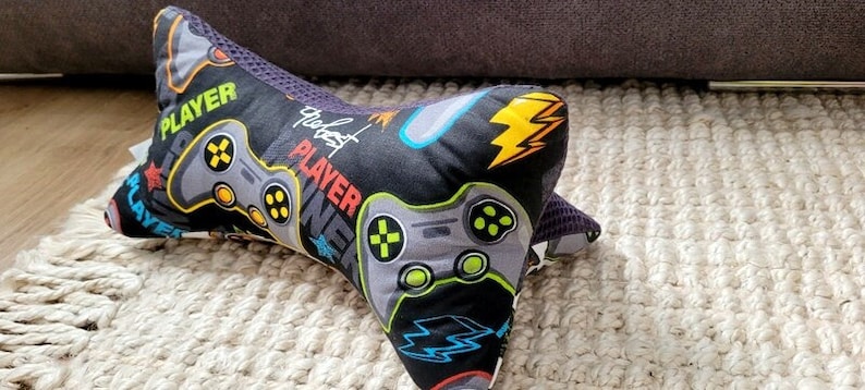 May include: A black and gray gaming pillow with a gray and green video game controller design. The pillow has the text "The Best Player" on it.